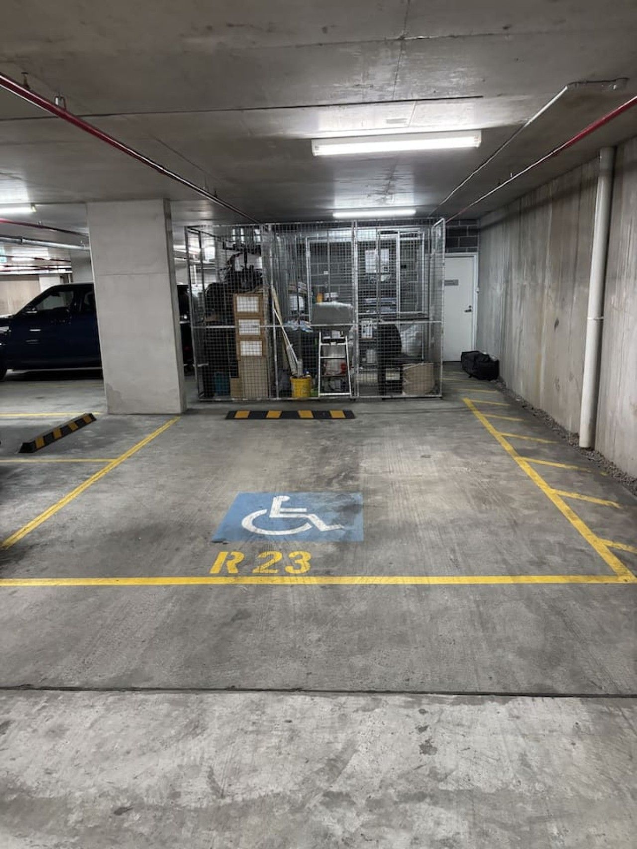 Un parking souterrain avec une place de stationnement réservée aux personnes handicapées, marquée R23, une barrière métallique et un symbole bleu représentant les personnes handicapées peint sur le sol.