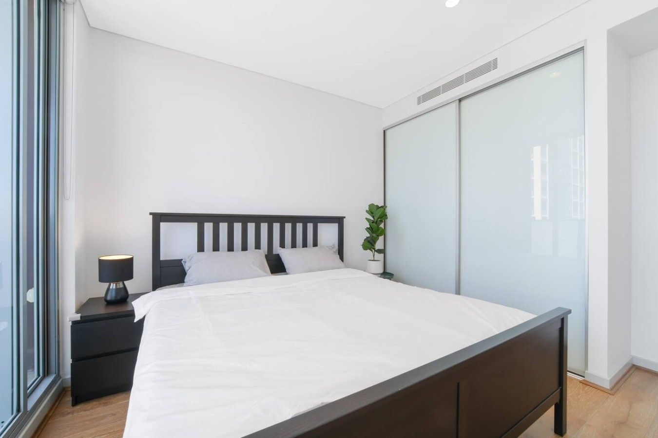 A modern bedroom with a large bed, a bedside table with a lamp, a potted plant, and a sliding closet door.