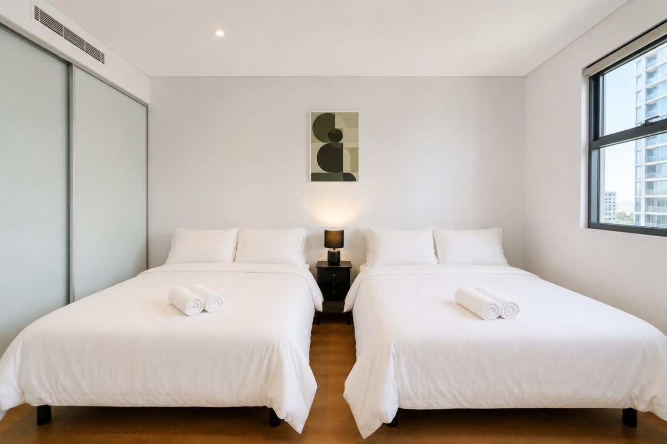 Een hotelkamer met twee bedden, een nachtkastje met lamp, een schilderij aan de muur en een raam met uitzicht op de stad.