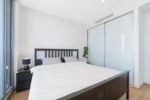 Een moderne slaapkamer met een groot bed, een nachtkastje met lamp, een potplant en een inloopkast met glazen schuifdeuren.