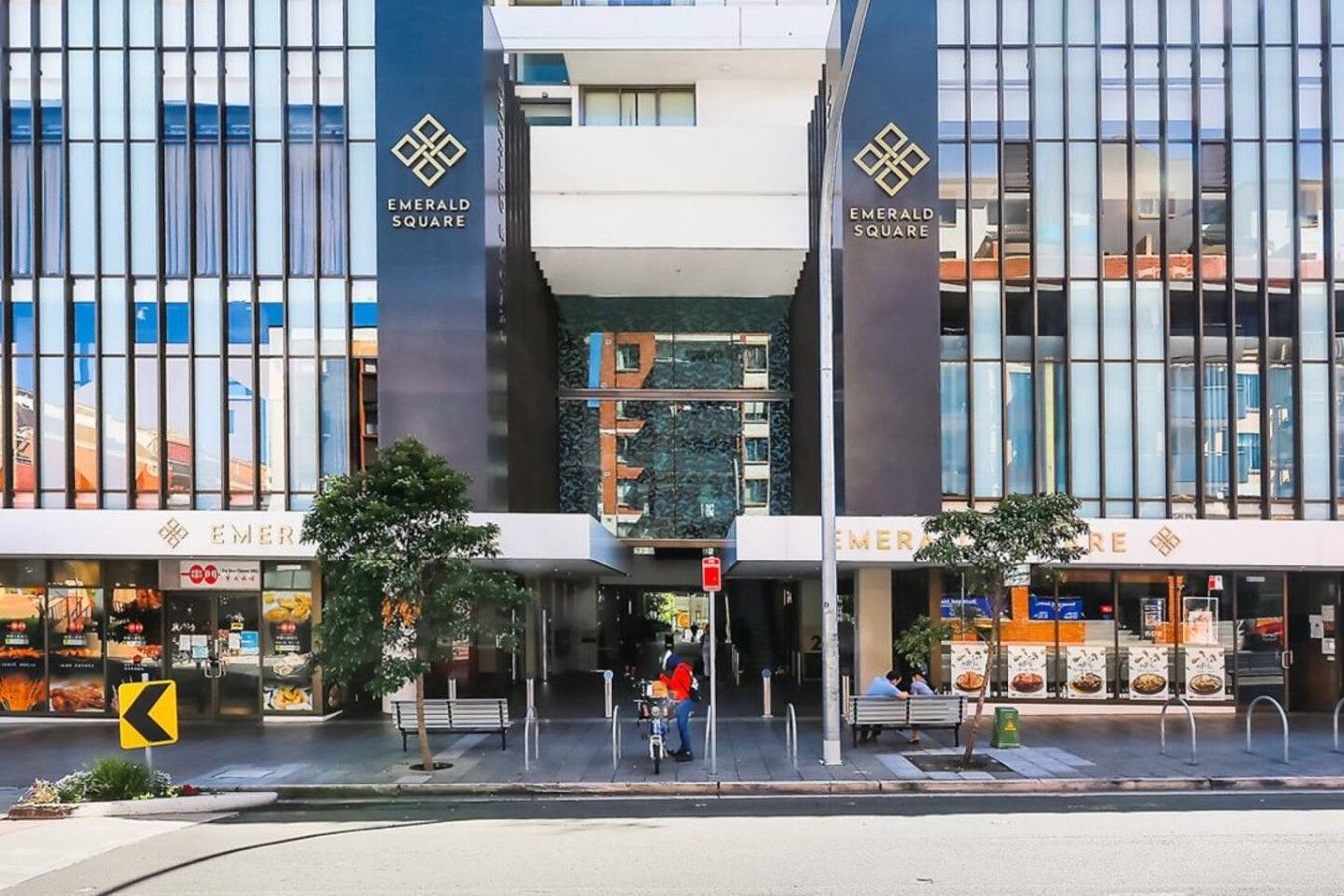 A modern building with 'Emerald Square' signage, featuring a central entrance with a glass facade, benches, and a pedestrian crossing.