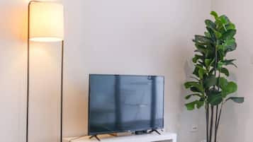 A flat-screen TV on a white stand against a wall, next to a tall floor lamp and a potted plant.