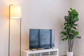A flat-screen TV on a white stand against a wall, next to a tall floor lamp and a potted plant.