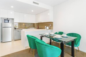 Cozinha moderna com armários brancos, uma ilha central e um espaço de refeições com mesa de vidro e cadeiras verdes.
