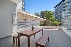 Um terraço no telhado com uma mesa e bancos de madeira, um vaso de planta e vista de um edifício ao fundo.