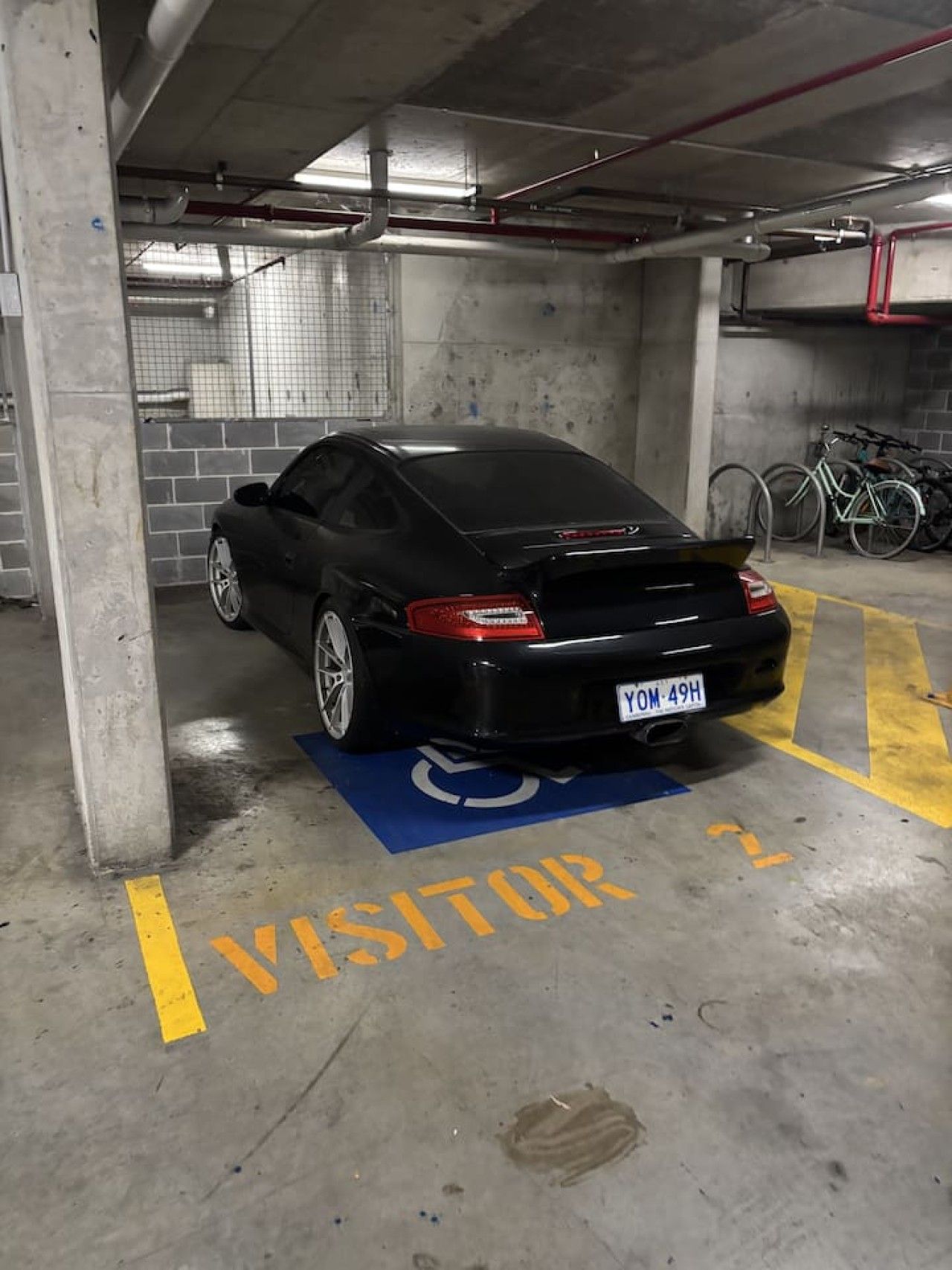 A black car parked in a visitor parking area of a building with concrete pillars and a tiled wall.