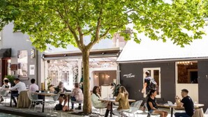 Een terras met tafels en stoelen, een boom die schaduw geeft, en een gebouw met een bordje op de achtergrond.