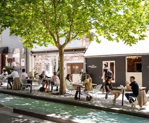 En udendørs café med borde og stole, et træ der giver skygge, og en bygning med et skilt i baggrunden.