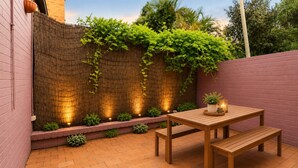 Een terras met een houten tafel en bank, een metselstenen vloer en een muur met klimplanten en verlichting.