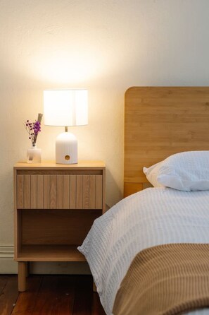 Ein ordentlich bezogenes Bett mit einem hölzernen Kopfteil, ein Nachttisch mit einer Lampe und eine Vase mit Blumen.