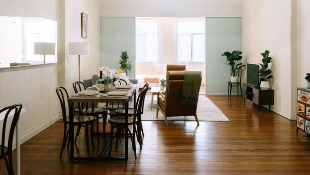 Een eetruimte met een houten tafel voor vier personen, omringd door stoelen. Achtergrond is een bank en een fauteuil, en een klein tafeltje met een plantje.