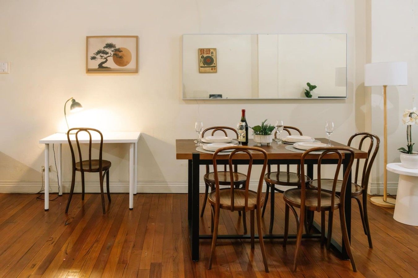 Un coin repas avec une table en bois dressée pour quatre personnes, entourée de chaises, un bureau blanc avec une lampe et des œuvres d’art accrochées aux murs.