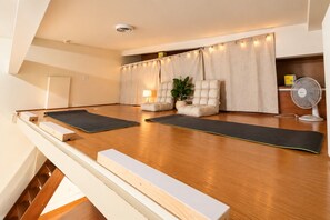 Een kamer met een houten vloer, twee yogamatten, een zachte bank, een potplant, een staande ventilator en een gordijn met sfeerverlichting.