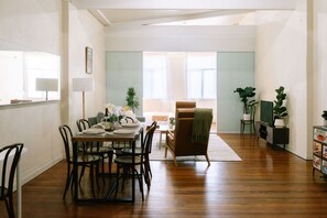 Een eethoek met een houten tafel gedekt voor vier personen, omringd door stoelen. Op de achtergrond staat een bank en een fauteuil, plus een kleine tafel met een lamp. De kamer heeft houten vloeren en uitzicht op het buiten via een raam.