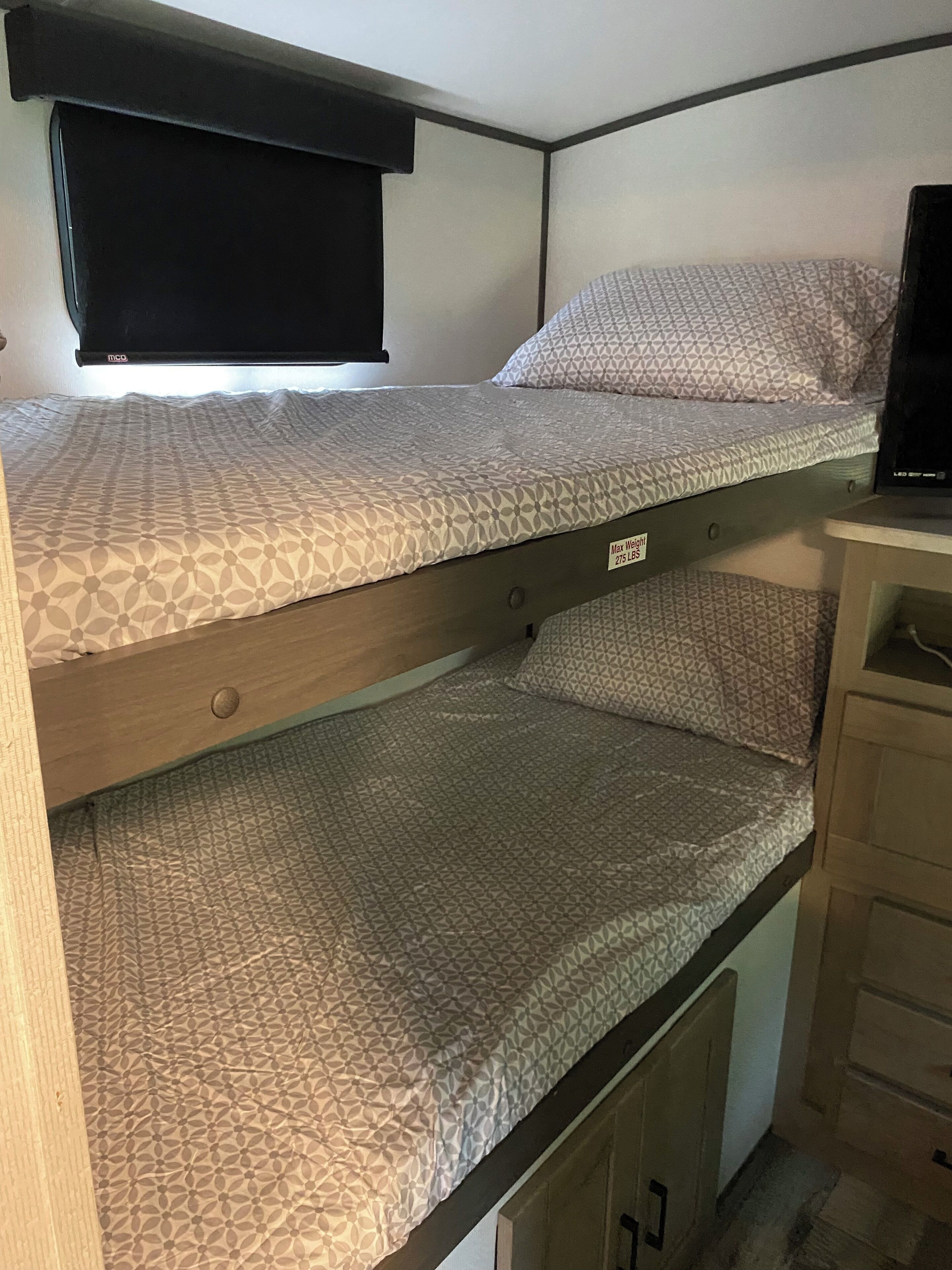 Ein Stockbett mit gemusterten Bettbezügen, ein kleiner Schreibtisch mit Computer und ein Fenster mit Jalousien.