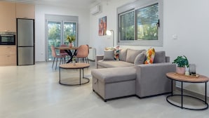 Una sala moderna con un sofá gris, una mesa de comedor con sillas, una zona de cocina con nevera y horno, y un ventanal con persianas.