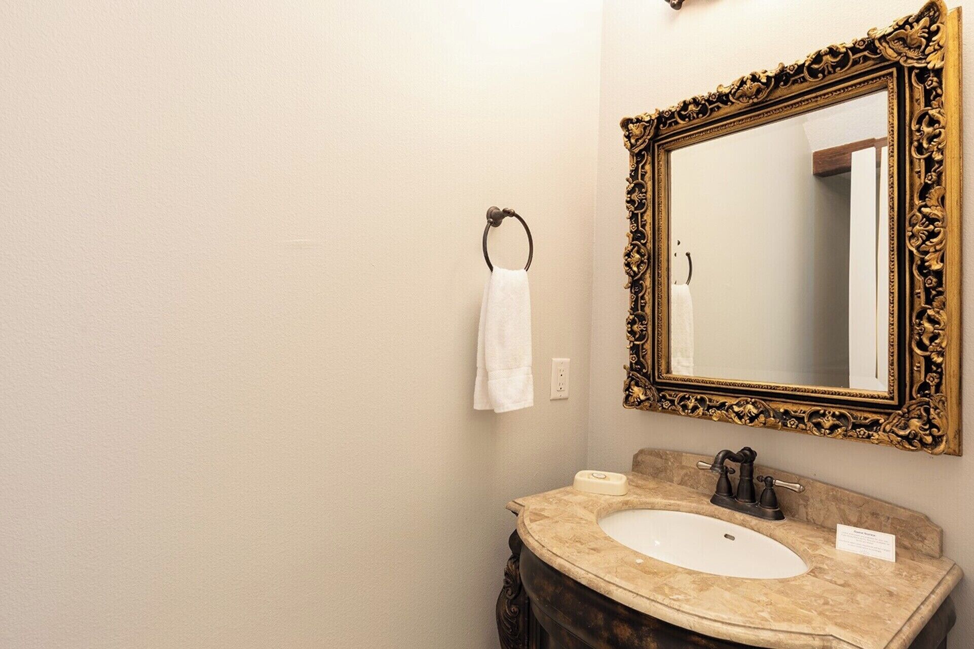A bathroom with a beige wall, a white sink, a gold-framed mirror, and a towel hanging on a metal ring.