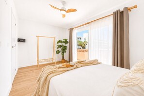 Ein Hotelzimmer mit Bett, Deckenventilator, Fenster mit Vorhängen, einer Pflanze und Blick auf einen Balkon.