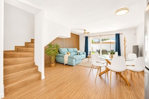 Ein modernes Wohnzimmer mit Treppe, einem blauen Sofa, einem Essbereich mit weißen Stühlen und einem großen Fenster.