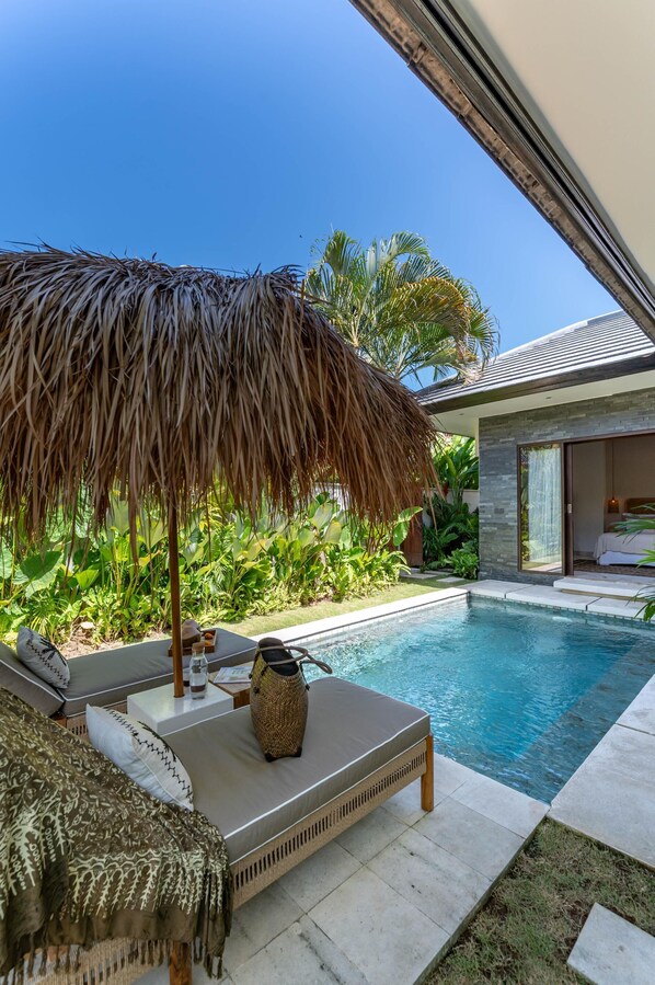 Un espace aménagé au bord de la piscine, avec un parasol en chaume, des chaises longues et une vue sur un bâtiment moderne.