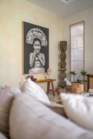 A room with a large framed black and white portrait, a wooden side table with a woven basket, a tall wooden sculpture, and potted plants. - Villa Athena One - Where Romance Meets Relaxation (Denpasar)