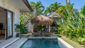 Piscina com guarda-sol de palha, cercada por plantas tropicais e uma parede de pedra.