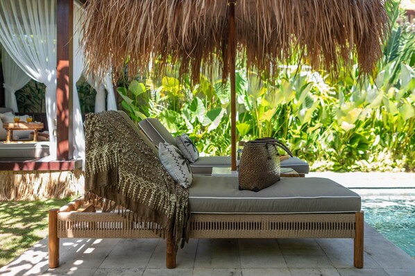 A wicker lounge chair with a cushion and a woven basket, positioned near a pool with lush greenery in the background. - Villa Athena One - Where Romance Meets Relaxation (Denpasar)