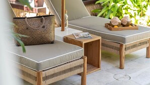 Uma área de estar à beira da piscina, com mobiliário de vime, um guarda-sol de palha e uma mesa de madeira com uma revista.