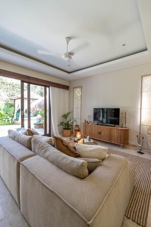 A modern living room with a large sofa, a TV on a wooden cabinet, and a ceiling fan. - Villa Athena 2 - Love, Leisure, and Bali Charm (Denpasar)