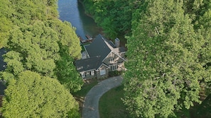 An aerial view of a house surrounded by dense green trees, with a visible driveway leading to the entrance.
