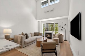 Ein geräumiges Wohnzimmer mit einer Sofagarnitur, einem Couchtisch, einem Kamin und einem großen Fenster mit Blick auf Bäume.