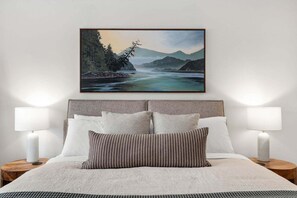 Ein Schlafzimmer mit einem Bett, zwei Nachttischlampen, einem Kopfteil und einem gerahmten Landschaftsbild über dem Bett.