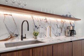 Eine Küche mit Marmorfliesen an der Spritzschürze, einem schwarzen Wasserhahn, einer Kaffeemaschine und einem Holzregal über dem Spülbecken.