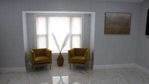 Een kamer met twee gele fauteuils, een groot raam met jaloezieën, een vaas met witte veren, en een schilderij aan de muur.