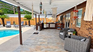 Un patio techado al aire libre con piscina, muebles de mimbre y una pared de ladrillo.