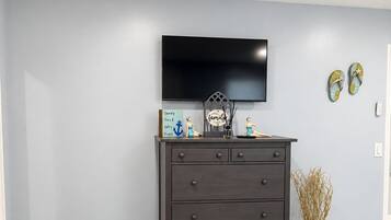 Quarto com cômoda de madeira escura, TV de tela plana na parede e vaso com plantas secas no chão.