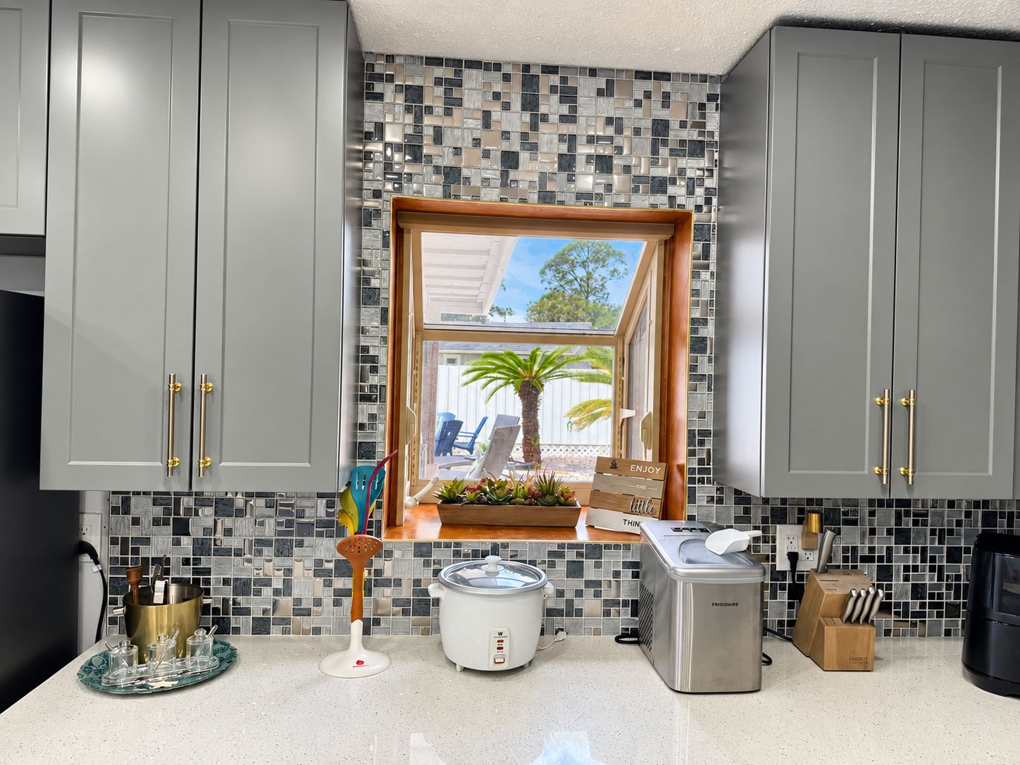 A kitchen with grey cabinets, a window with a view of palm trees, a crockpot, and a toaster.
