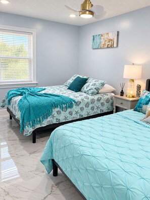 A bedroom with two beds, a ceiling fan, a window with blinds, a nightstand with a lamp, and a wall art piece. - Enjoyable 3-bedroom house with pool and AC in charming Jacksonville, Florida. (Jacksonville)