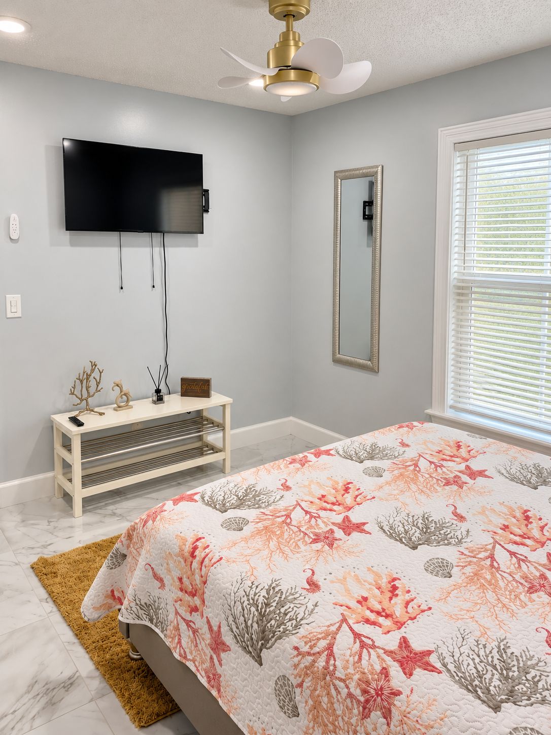 A bedroom with a bed, a TV mounted on the wall, a bedside table, and a window with blinds.
