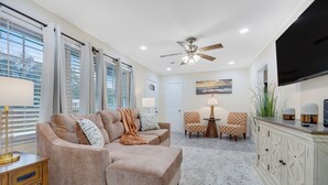 Un salon avec un canapé d’angle beige, une table basse, deux fauteuils à motifs, un téléviseur et un ventilateur de plafond.