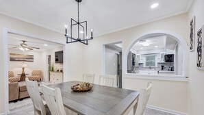 Un coin repas avec une table en bois, des chaises blanches et un lustre. On peut apercevoir la cuisine à travers une arche.