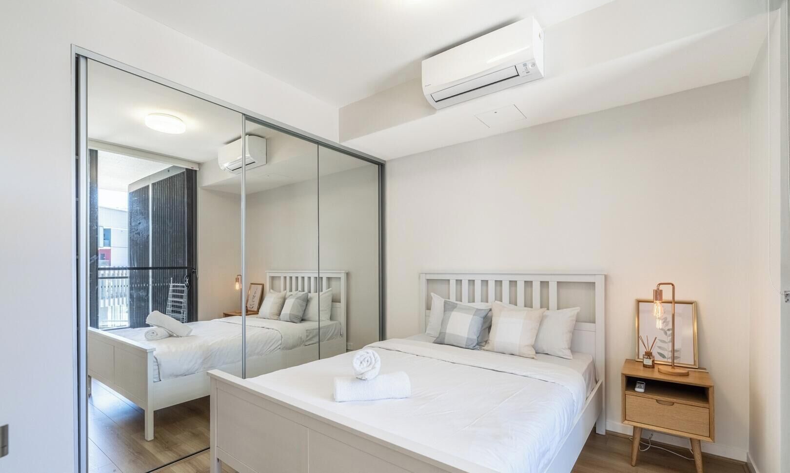 Un dormitorio con un espejo grande, una cama con ropa blanca, una mesita de noche con lámpara y un equipo de aire acondicionado.