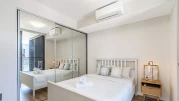 Un dormitorio con un espejo grande, una cama con ropa blanca, una mesita de noche con lámpara y un equipo de aire acondicionado.