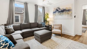 Una sala de estar con un sofá seccional, un sillón, una mesita con lámpara y un cuadro en la pared.