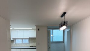 A spacious, well-lit room with a large window, a kitchen area in the background, and a wooden floor. - Happy house (Yongin-si)