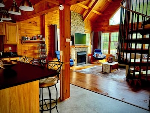 A wooden cabin interior with a kitchen, dining area, and living room, featuring a fireplace and large windows.