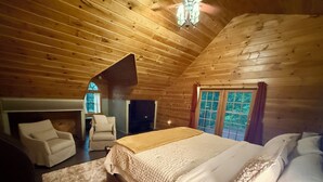 Een slaapkamer in een houten chalet, met een bed, twee stoelen, een open haard en een raam met gordijnen.