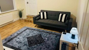 A living room with a dark grey sofa, a patterned rug, a small table with a lamp, and a white door. - Modern Apartment 5 Minutes from City Airport (London)