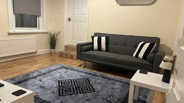 A living room with a grey sofa, a patterned rug, a white side table, and a window with a blind. - Modern Apartment 5 Minutes from City Airport (London)