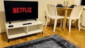 A living room with a TV displaying the Netflix logo, a white entertainment unit, a dining table with chairs, and a patterned rug. - Modern Apartment 5 Minutes from City Airport (London)